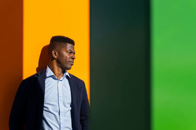 African american male in formal wear frowning face from sunshine while leaning on bright wall and looking away