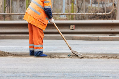 A road service worker in a bright orange reflective uniform shovels loose sand with a shovel .