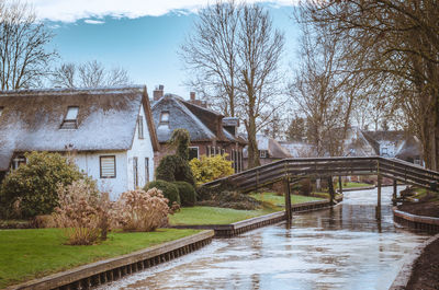 Bridge over river amidst bare trees and buildings against sky