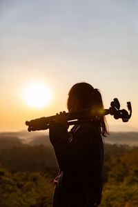 Side view of girl standing with tripod against sky during sunset