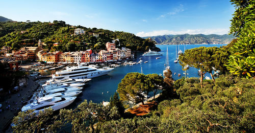 Yachts moored in sea at harbor by village against blue sky