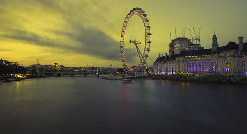 Illuminated ferris wheel by river against sky in city