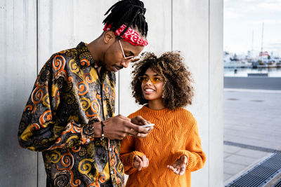 Cheerful black woman with curly hair standing with cool ethnic guy in sunglasses and smiling while using mobile phone against gray wall in city