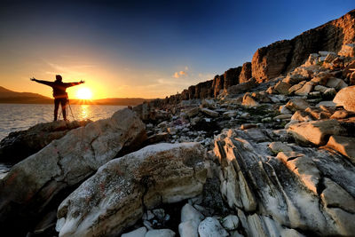 Rear view of silhouette man with arms outstretched standing on rock at beach during sunset