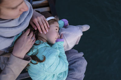 High angle view of daughter lying on mother's lap while sitting on pier over sea