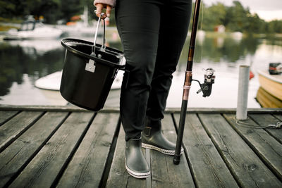 Low section of woman with fishing rod and basket standing on pier near lake