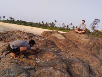 Man photographing friend sitting in lotus position on rock formation at beach against sky