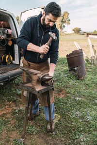Worker in uniform forging horseshoe with hammer on metal anvil while working in rural area on summer day