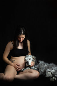 Portrait of pregnant woman with dog sitting against black background