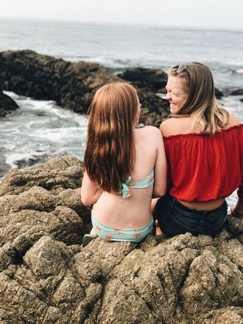 REAR VIEW OF WOMEN SITTING ON ROCK BY SEA