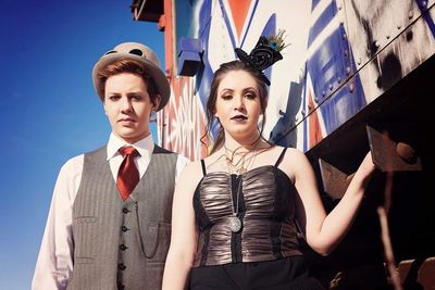 Low angle portrait of couple standing by metallic structure on sunny day