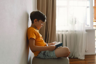 A cute boy wearing an orange t-shirt is sitting on a soft ottoman reading a book