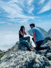 Man proposing woman while siting on mountain against sky