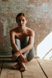 Portrait of shirtless young woman sitting on floor against wall