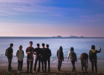 People standing on shore at beach against sky during sunset