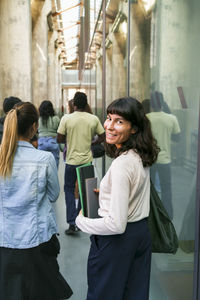 Smiling female student looking back while walking with friend in college corridor