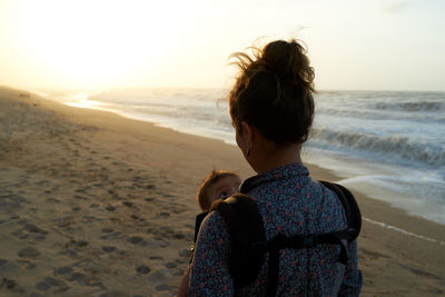 Rear view of woman carrying son at beach against sky during sunset