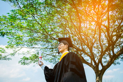 Low angle view of man in graduation gown standing against tree