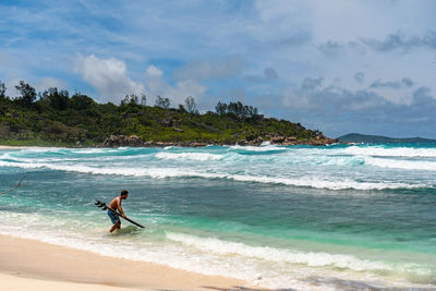 Man wearing swim trunks, standing on paradise beach, going surfing in turquoise ocean with waves