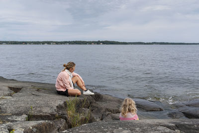 Woman and young girl sitting on rock by sea against sky