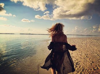 Rear view of sensuous young woman running at beach against sky