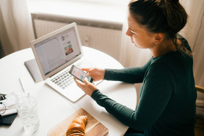 Woman using smart phone while surfing food recipe on laptop at home