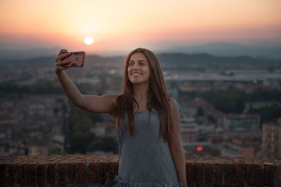 Smiling young woman using phone against sky during sunset