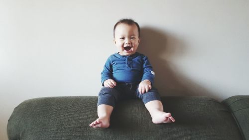 Portrait of cute baby boy with mouth open sitting on sofa against wall at home