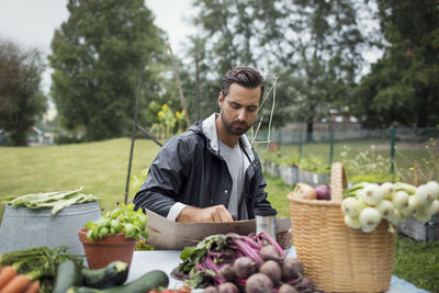 Mid adult man writing on cardboard while sitting by freshly harvested vegetables in urban garden