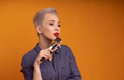 Thoughtful young woman with make-up brushes standing against orange background