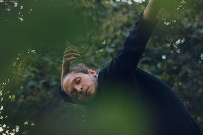 Thoughtful young man dancing with eyes closed against trees 