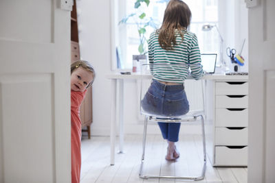 Girl standing behind door while mother working on laptop in background at home