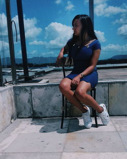 Young woman looking away while sitting on chair against blue sky during sunny day