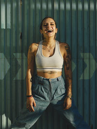 Happy young female with tattoos wearing stylish outfit standing near wall in city and looking at camera