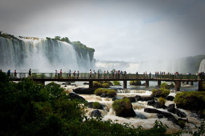 Group of people on footbridge over river by waterfalls against the sky