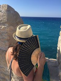 Rear view of woman wearing hat while holding hand fan by sea 