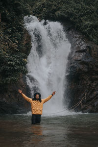 Portrait of man with arms raised standing against waterfall