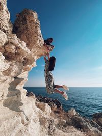 Full length of woman holding hand mid-air by rock formation against sky and sea