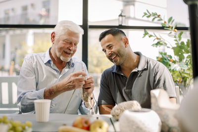 Happy male nurse sitting with senior man crocheting at nursing home