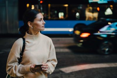 Woman with cellphone looking away while standing in city at night