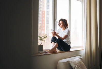 Young brunette woman in casual clothes using mobile phone sitting on window sill at home