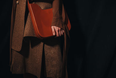 Female figure dressed in stylish beige outfit and holding brown leather bag on black background
