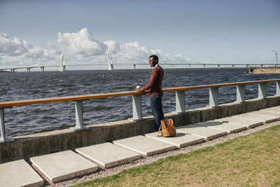 Young man sitting on railing at the sea, eating hamburger, drinking coffee