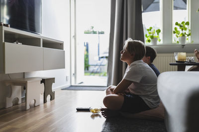 Siblings watching tv while sitting on carpet in living room at home