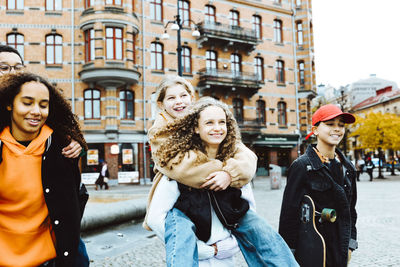 Happy girls giving piggyback ride to male and female friends on footpath in city