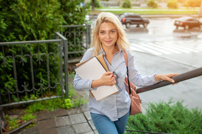Portrait of woman with books standing by railing in city