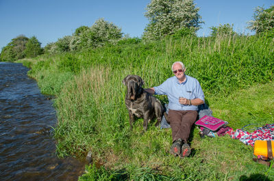 Full length of senior man with neapolitan mastiff on grassy field by river