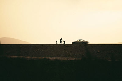 Silhouette of man and boys standing by car against sky during sunset