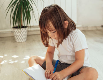 Attentive child sits on the floor at home with a laptop, pen and notebook. 