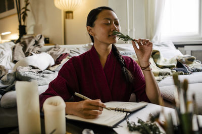 Young woman smelling herbs while while writing in book at home